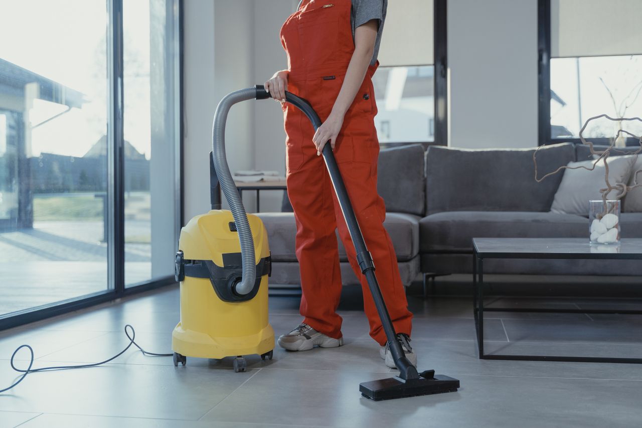 Un nettoyeur professionnel en combinaison rouge aspirant un salon moderne avec un aspirateur jaune.